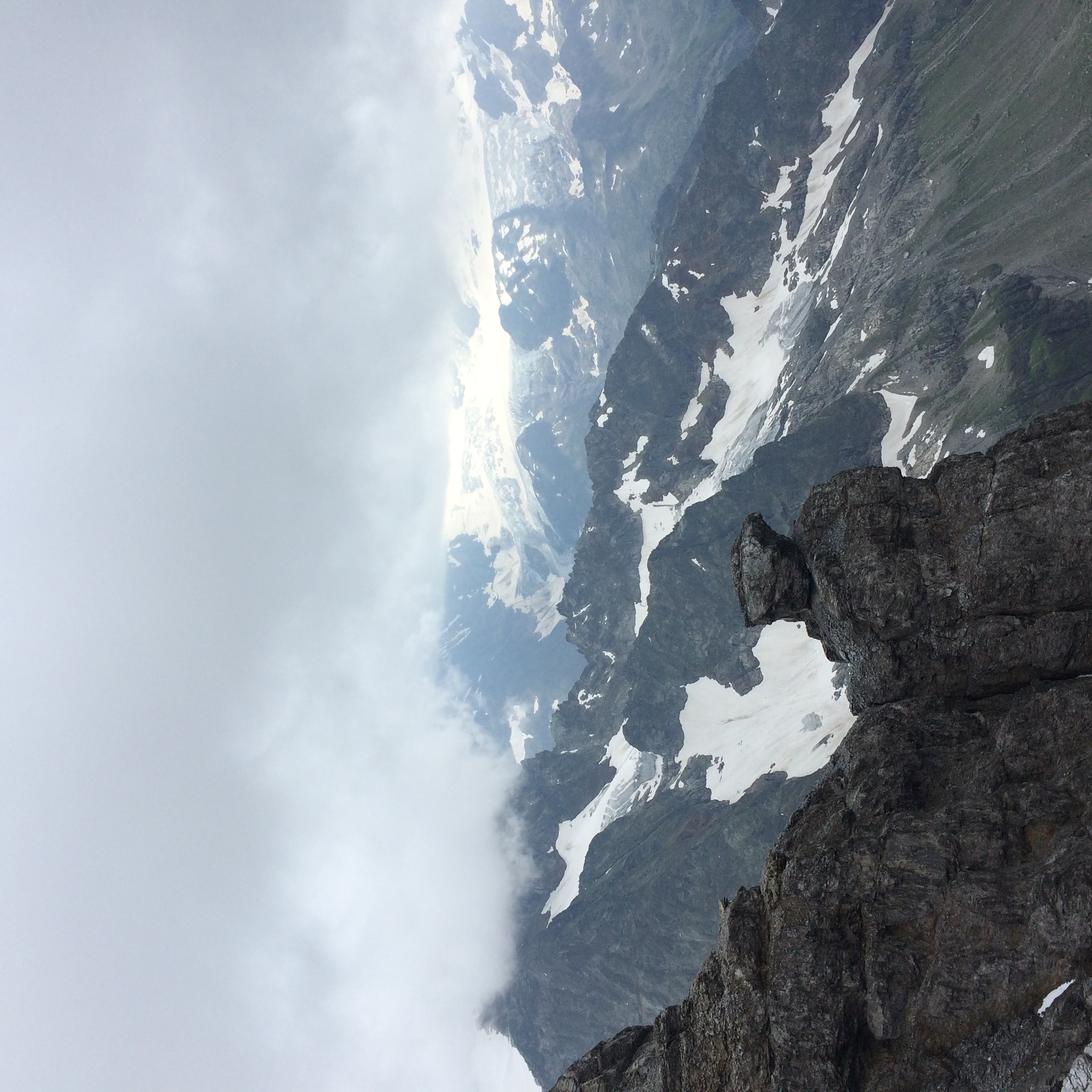 Clouds cover the peaks of rugged, rocky mountains with patches of snow and ice. A distinctive rock formation stands in the foreground, with valleys and green slopes visible below.