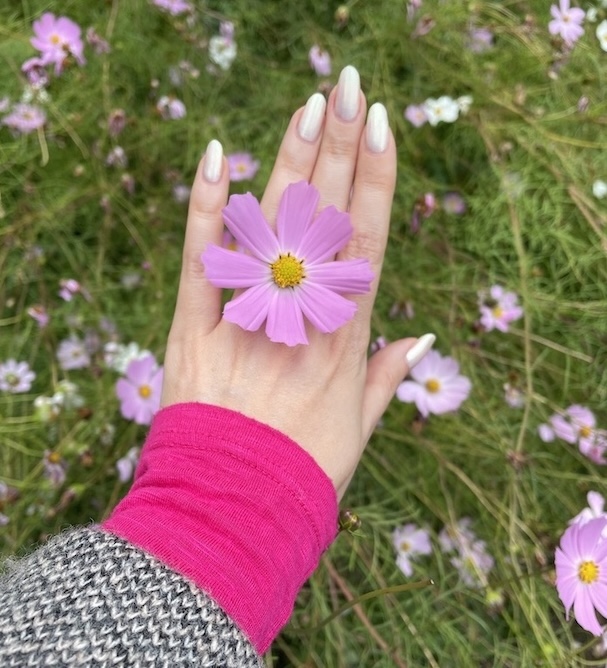A beautiful gentle woman's hand with light-colored nails holds a pink cosmos flower over a field of similar flowers. The person is wearing a pink sleeve under a black-and-white patterned coat.