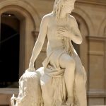 A beautiful statue of Amalthea in marble beside the sea goat that fed Zeus milk. The statue is beautifully lit by the sun and shows the woman in a gentle sitting pose touching her chest and looking at the horizon.