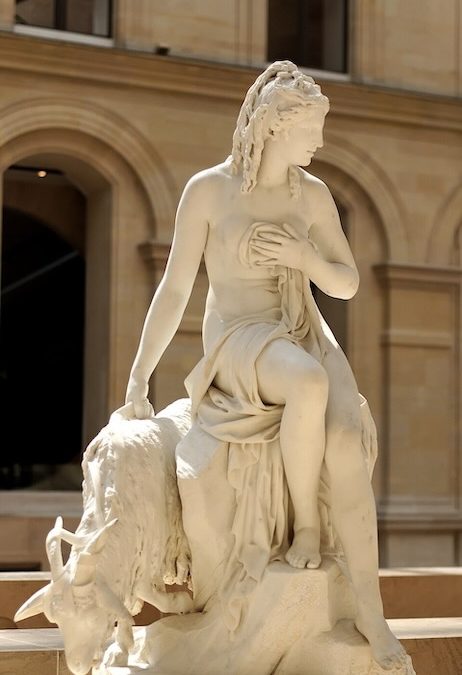 A beautiful statue of Amalthea in marble beside the sea goat that fed Zeus milk. The statue is beautifully lit by the sun and shows the woman in a gentle sitting pose touching her chest and looking at the horizon.