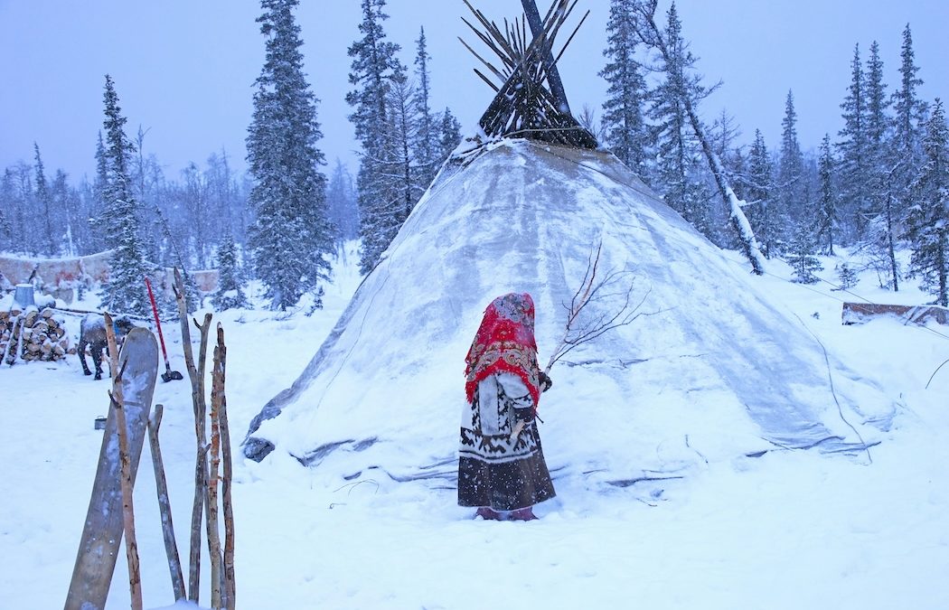 A girl from the Nenets Indigenous people of Siberia standing outside their tent in winter, with a reindeer in the distance.
